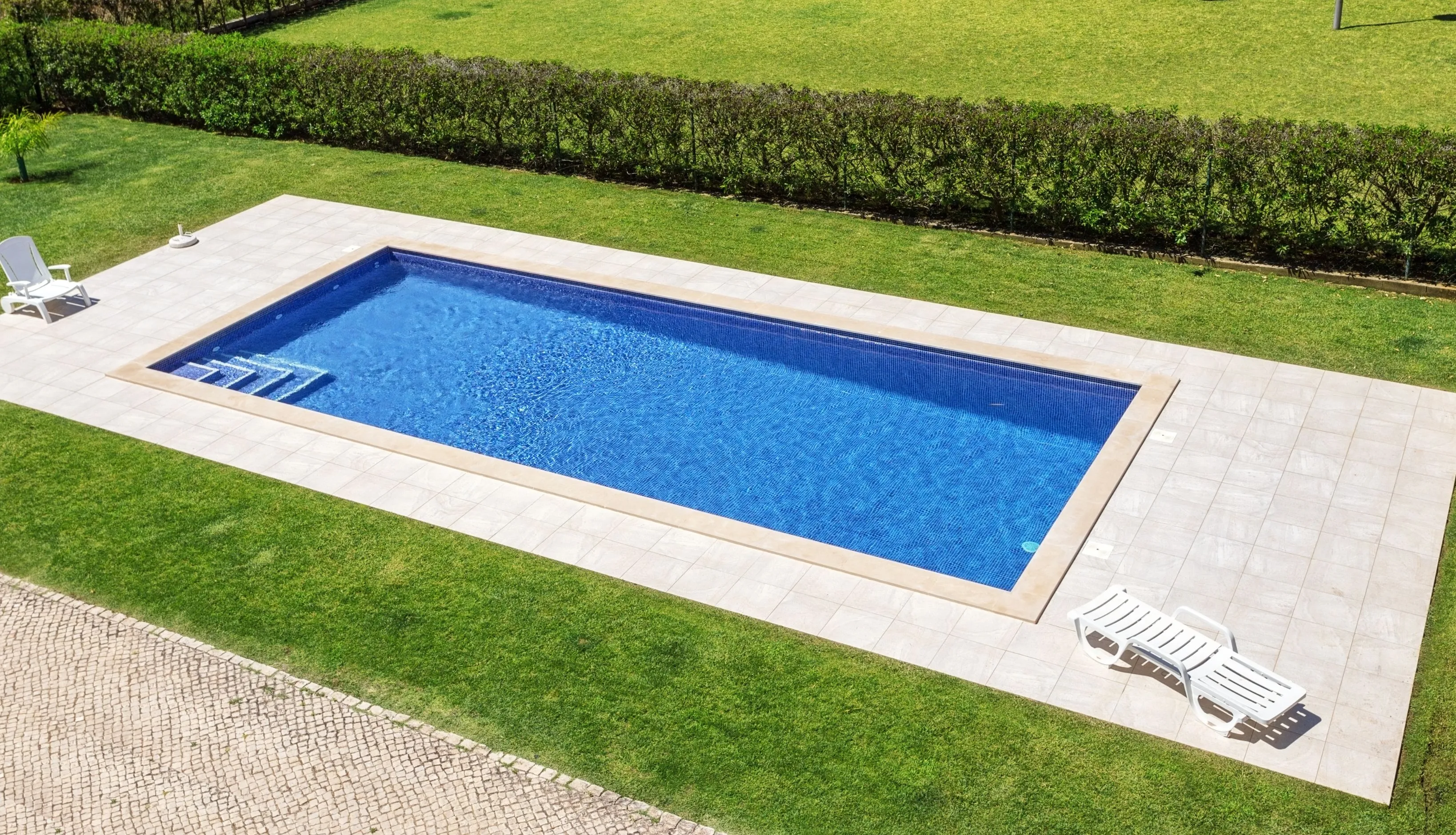 The stunning result of a professional pool remodeling in Los Angeles featuring deep blue mosaic tiling, smooth light-colored stone pavers, and crystal clear water.